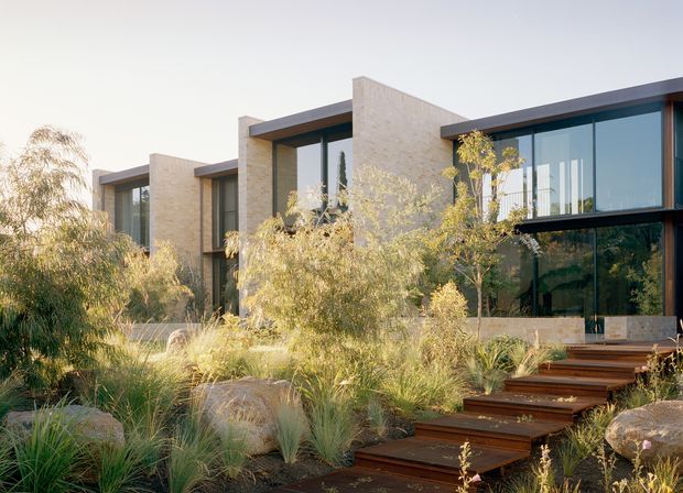 A contemporary low-profile residence set within a landscaped garden, featuring rectilinear volumes, large floor-to-ceiling glazed openings, dark roof edges and frames, and pale textured masonry walls. Timber stepping stairs lead through ornamental grasses, shrubs, and large natural boulders toward the entrance.
