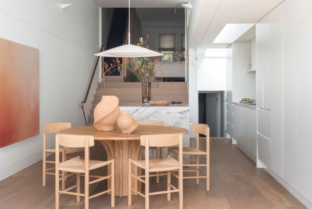 Bright contemporary open-plan dining and kitchen interior with a sculptural light-wood dining table and matching ladder-back chairs in the foreground. A marble-clad island separates the dining area from the kitchen, which features streamlined white floor-to-ceiling cabinetry and integrated appliances. A staircase, timber-panelled wall, indoor planting, skylights, and a large suspended pendant add architectural interest. The palette is light, warm, and refined.