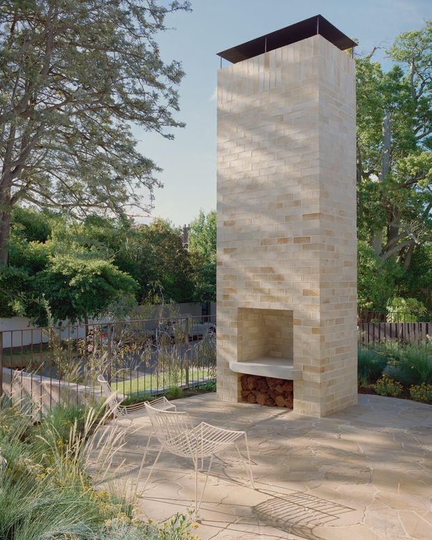 A landscaped outdoor patio featuring a tall freestanding masonry fireplace with a broad dark metal cap. The fireplace is built from pale rectangular stone or concrete blocks and has an open firebox with stacked firewood. Light-toned irregular stone paving, wire lounge chairs, ornamental grasses, mature trees, planted garden beds, and a metal railing surround the outdoor entertaining area.