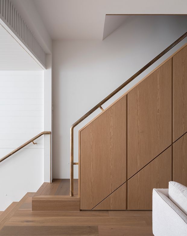 A refined interior stairway with warm timber flooring and a full-height timber-clad stair balustrade or storage wall. Slim bronze-toned handrails trace the staircase, while white walls, a white horizontal-panel door, and a minimalist ceiling opening create a clean, contemporary composition. The corner of a light upholstered sofa is visible at lower right.