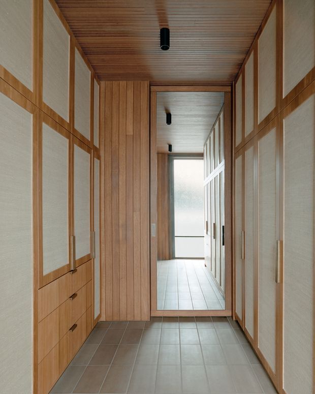 A narrow, highly tailored hallway lined with floor-to-ceiling timber cabinetry and framed light fabric or textured panels. The ceiling is clad in closely spaced timber slats with simple black cylindrical downlights. Large rectangular grey floor tiles lead toward a bright glazed doorway or window, while a full-height mirror at the end visually extends the corridor. The scheme is warm, restrained, and strongly defined by repeated timber framing and integrated storage.