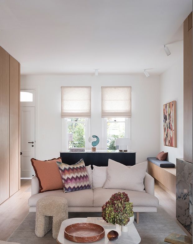 Bright, compact contemporary living room with a neutral palette, pale timber built-in cabinetry, white walls, a low upholstered sofa, and a marble-topped coffee table. Two windows with fabric Roman blinds provide natural light, while a dark built-in console, sculptural decor, artwork, and patterned cushions add contrast and colour. A concealed entry door and integrated cabinetry are visible at the left and right edges.