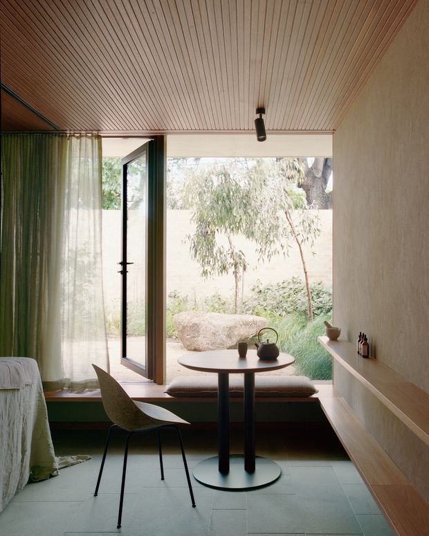 A softly lit compact dining nook beside a full-height glazed door and window, with a small round table, two dark pedestal legs, sculptural chairs, and a built-in window bench. The room features a warm narrow timber-slat ceiling, textured pale plaster walls, a long built-in timber shelf and cabinetry, and large muted green floor tiles. Sheer curtains frame a view to a planted courtyard, creating a calm, intimate interior with strong indoor-outdoor connection.