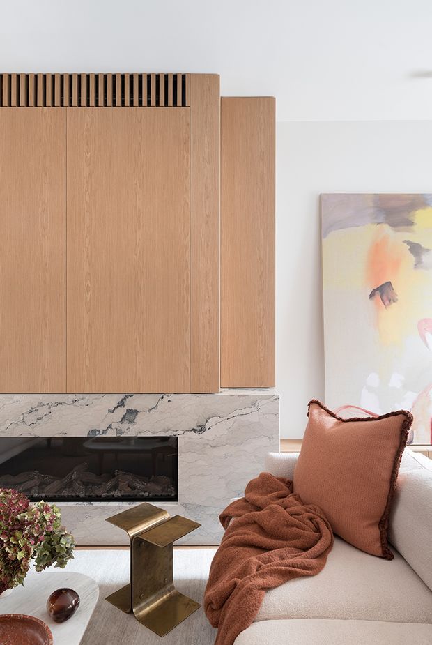 Close-up of a refined contemporary living room featuring a built-in timber fireplace wall with vertical ventilation slats, a pale veined stone fireplace surround, a light upholstered sofa, and warm rust-toned cushions and throw. An abstract artwork and small metallic side table add understated decorative detail.