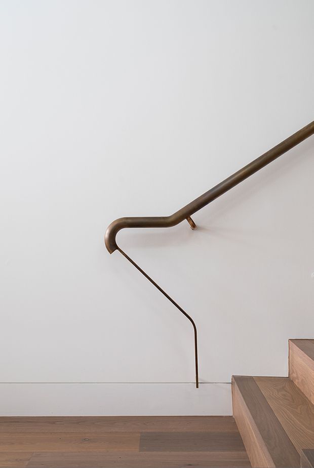 Close-up architectural detail of a minimalist staircase with a slender bronze-toned metal handrail and matching thin balustrade element mounted against a smooth white wall. The stair treads and surrounding floor are finished in warm natural timber, creating a clean, refined contemporary composition.