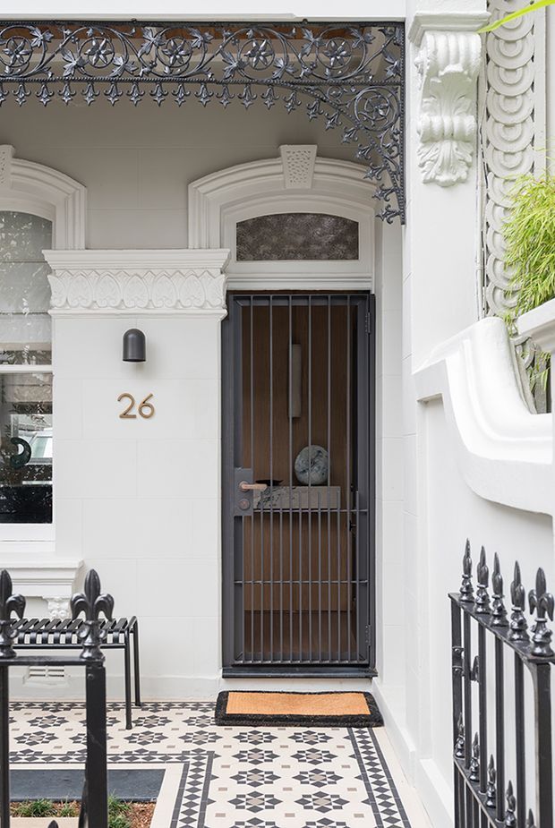 A refined heritage-style residential entryway viewed from outside, featuring ornate white-painted architectural detailing, decorative wrought-iron canopy and gate, a dark timber front door, patterned geometric encaustic-style paving, and the house number 26. The facade includes carved mouldings, arched window surrounds, a projecting pilaster-like entry element, and black exterior wall lighting.