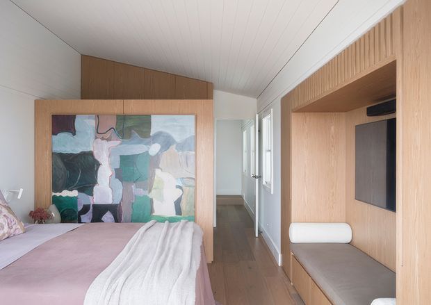 A softly styled bedroom with a partially visible bed, pale mauve bedding, and a large abstract artwork framed within warm timber joinery. The room features white vertical-panel ceilings and walls, timber flooring, a built-in media and seating wall with a wall-mounted screen, and a narrow hallway leading to a bright adjoining area. The overall design is compact, warm, and contemporary with subtle mid-century influences.