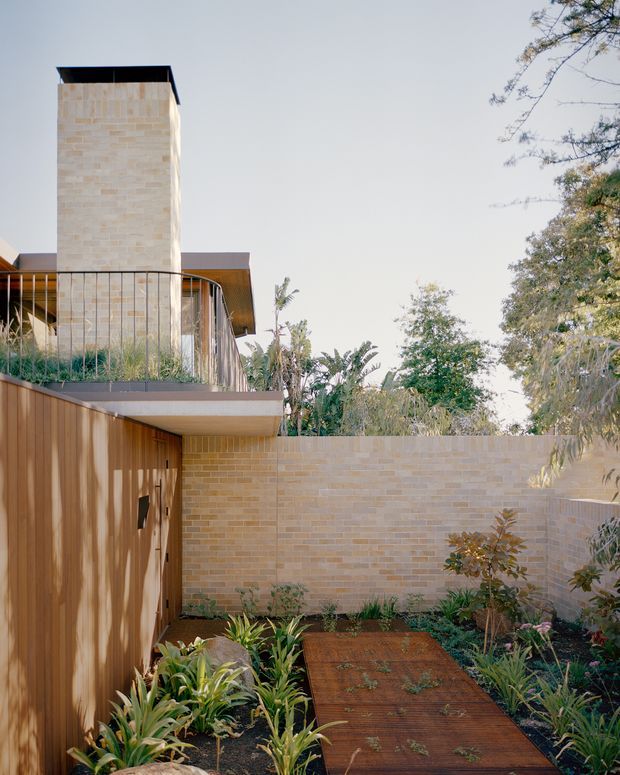A contemporary residential exterior showing a landscaped courtyard or garden path. Pale tan brick walls and a tall brick-clad tower sit above timber-clad lower walls, with a curved balcony railing and deep roof overhangs. A rust-toned metal walkway passes through dense planting toward the building, creating a warm, integrated indoor-outdoor setting.
