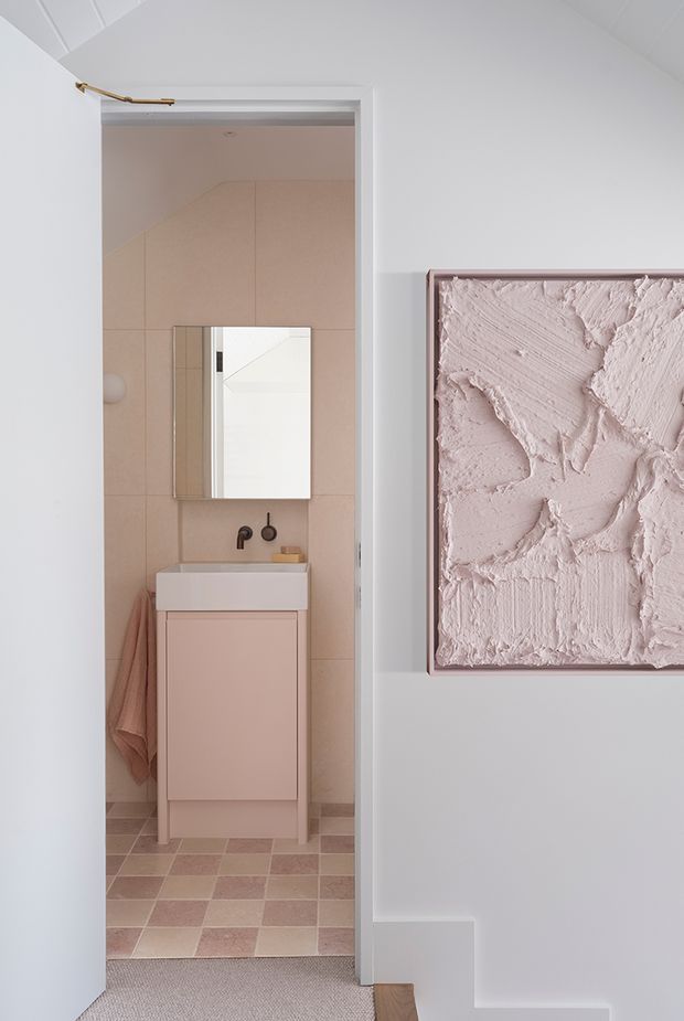 A softly styled view from a pale hallway into a compact bathroom. The bathroom has blush-pink tiled walls, a checkerboard pink and cream tiled floor, a wall-mounted white basin on a dusty-pink vanity, dark metal tapware, and a simple rectangular mirror. A textured blush artwork in a thin frame hangs prominently on the hallway wall. White painted walls and ceiling create a calm, refined interior.