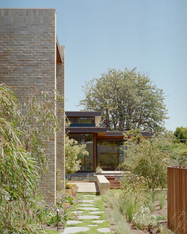 A landscaped residential courtyard viewed from an outdoor garden path. The scene features a contemporary house with dark brick masonry, extensive floor-to-ceiling glazing, slim dark frames, and low-pitched roof planes. Layered planting, ornamental grasses, stepping stones, timber decking, and a timber privacy screen create a natural, carefully designed garden setting.