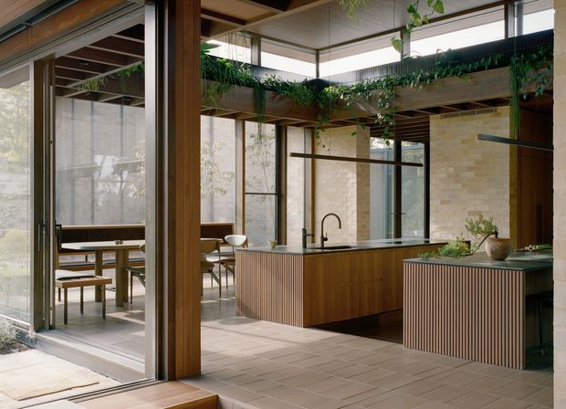 A refined contemporary kitchen and dining space with warm timber columns, fluted timber island cabinetry, pale tiled flooring, exposed structural beams, and extensive floor-to-ceiling glazing. Indoor planting runs along the upper beams and ledges, while a dining area is visible through the open glazed partition. The kitchen includes a dark benchtop, integrated sink and tap, and a separate curved island or storage unit.