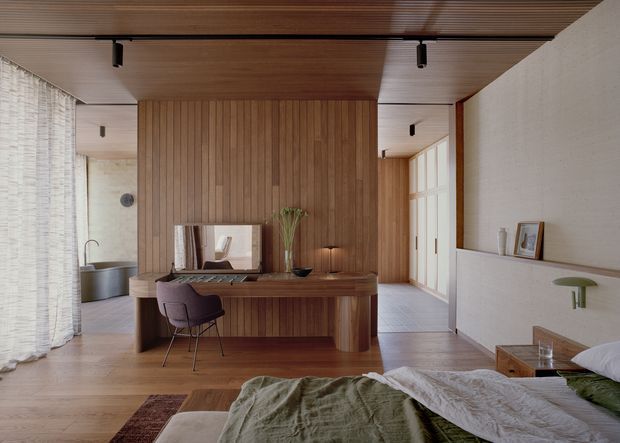 Warm, refined bedroom interior with a timber-lined ceiling and feature wall, integrated desk and vanity, upholstered chair, platform bed, and a partially visible freestanding bathtub behind sheer curtains. Natural light enters through full-height glazing and a glazed door, creating a calm contemporary retreat.
