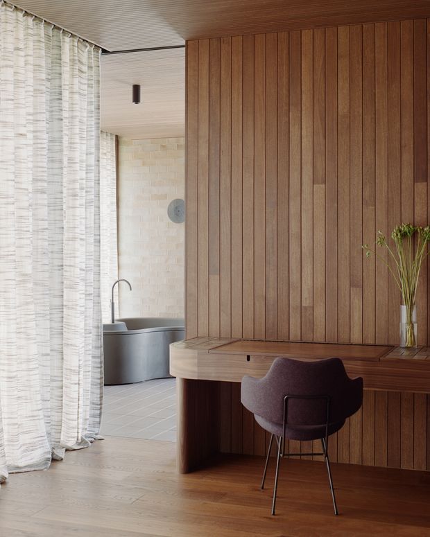 A warm, refined interior showing a built-in timber desk and cabinetry integrated into a full-height vertical wood-panelled wall. A dark upholstered chair sits at the desk, with a vase of dried stems on the right. Sheer textured curtains frame an adjoining bathroom containing a freestanding bath, pale brick wall finish, and tiled floor. The room has timber flooring and a timber-lined ceiling, creating a cohesive natural-material scheme.