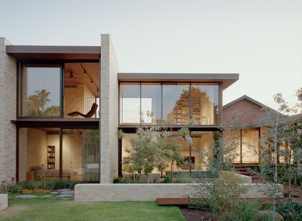 Contemporary multi-level residential facade with extensive floor-to-ceiling glazing, pale masonry walls, projecting dark roof planes, warm timber soffits, and landscaped courtyard planting. The composition combines a crisp modern addition with a visible pitched-roof brick structure behind it.