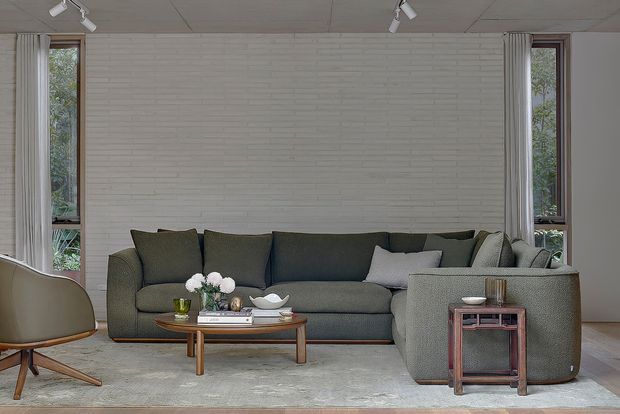 A calm, contemporary living room with a large dark grey sectional sofa, matching armchair, low timber coffee table, and a small side table. The room features a white painted brick feature wall, tall narrow windows with light curtains, pale timber flooring, a neutral rug, and track lighting.