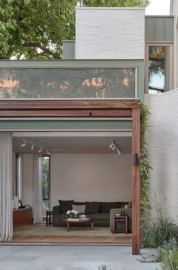 A refined residential facade with a large open glazed living-room frontage framed in warm timber. The exterior combines pale painted brick, muted green-grey metal or painted panels, expansive glazing, and a curved white-brick wall. Through the opening, a furnished living room with a dark sofa, timber coffee table, track lighting, curtains, and pale brick walls is visible. Landscaping and climbing greenery soften the contemporary composition.
