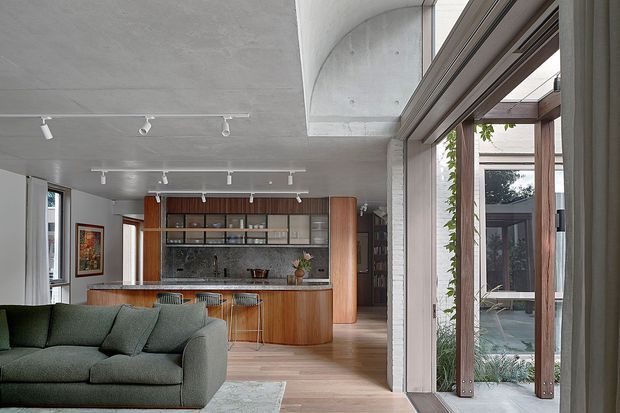 Open-plan contemporary living and kitchen interior with a deep green sectional sofa in the foreground, warm timber kitchen cabinetry and curved island, integrated shelving, pale timber flooring, exposed concrete ceiling and column, track lighting, and large glazed sliding doors opening to a planted courtyard. The room has a refined, warm modern character with strong indoor-outdoor connection.