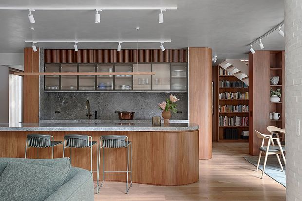 A refined open-plan kitchen and living area viewed from the lounge. The kitchen features a large curved timber island with a stone benchtop, three metal bar stools, dark stone splashback, timber cabinetry, and glazed upper cupboards. Track lighting runs across the ceiling, while a built-in timber bookcase and dining nook are visible beyond the kitchen. A pale upholstered sofa occupies the foreground, and a white-painted brick wall frames the right side.