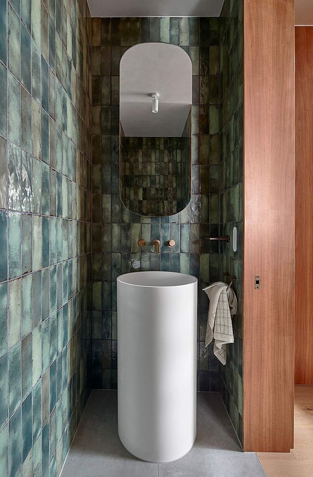 A narrow, highly detailed bathroom with full-height dark teal and green glazed tile walls, a tall cylindrical white pedestal basin, wall-mounted brushed metal tapware, and a rounded rectangular mirrored cabinet above. A timber-framed doorway and warm timber paneling contrast with the moody tiled enclosure. The floor is a smooth grey finish, and a towel hangs from a wall-mounted hook.
