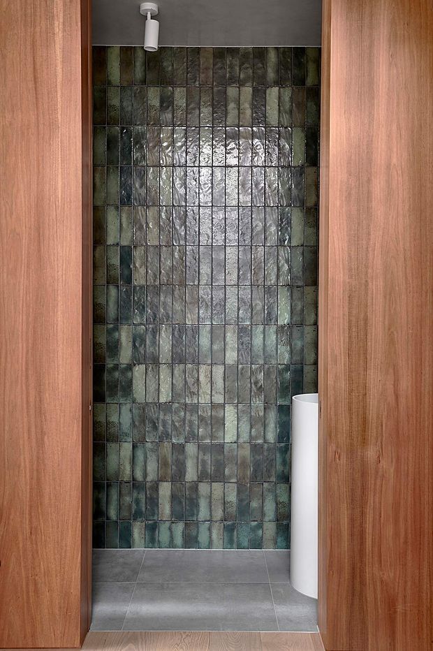 A narrow contemporary bathroom viewed through an open timber doorway. The room features a full-height wall of vertically arranged, irregularly textured glazed tiles in dark green, grey, and muted blue tones, a grey tiled floor, a white cylindrical wall-mounted fixture or basin edge, and a small white ceiling spotlight. Warm timber door panels frame the space.