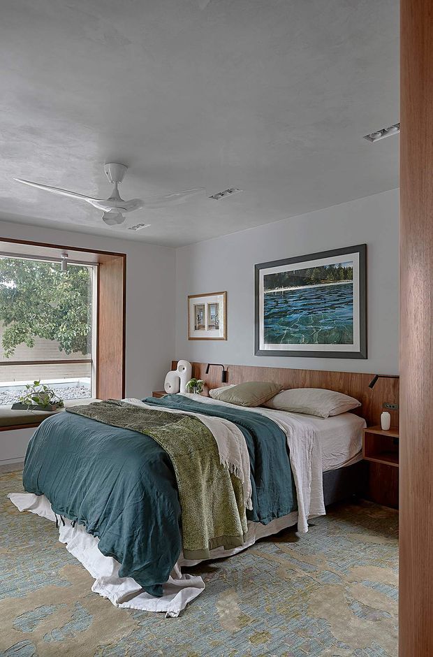 A contemporary bedroom with a low-profile bed, layered teal and olive bedding, and a timber feature wall behind the headboard. The room has pale textured walls and ceiling, recessed downlights, a ceiling fan, warm timber window and door framing, and large artwork above the bed. A broad window provides views of greenery, while patterned carpet covers the floor.
