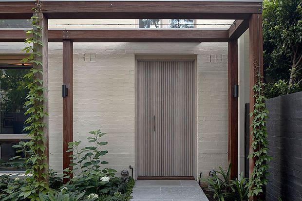 A refined garden entry framed by a dark timber pergola with translucent overhead panels and climbing greenery. The focal point is a tall vertically ribbed timber door set into a pale painted brick wall, approached by a grey stone-paved path and surrounded by dense planting. A dark masonry boundary wall and glimpses of glazing are visible at the sides.