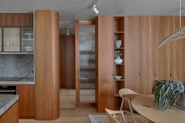 A warm, wood-lined open-plan kitchen and dining area with extensive built-in cabinetry. The image shows a stone-look kitchen benchtop and splashback at left, tall curved timber cabinetry, a glazed internal storage or pantry door, open display shelving, and a round timber dining table with sculptural wooden chairs. The room has a smooth grey ceiling, timber flooring, restrained contemporary detailing, and abundant natural wood tones.