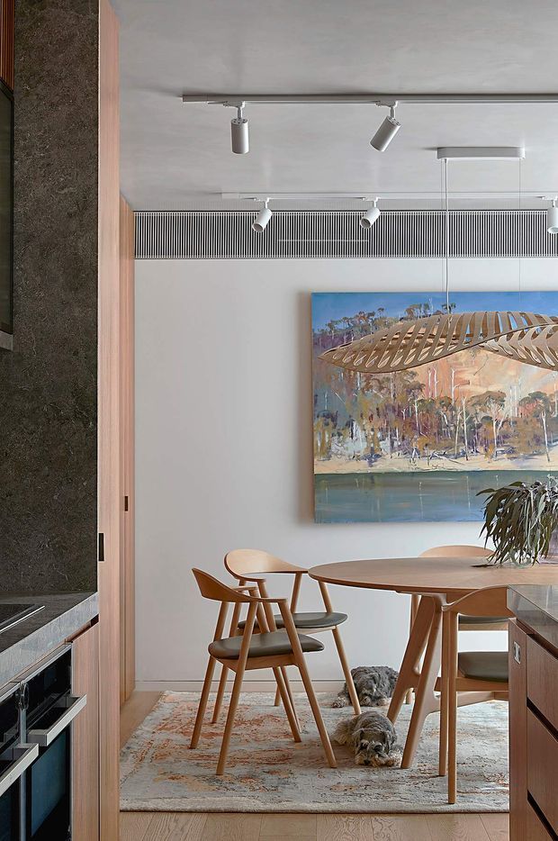 A refined contemporary dining area viewed from the kitchen, with a round timber dining table and sculptural wooden chairs on a patterned rug. A large Australian landscape artwork anchors the pale wall, while a linear pendant light and adjustable track lighting illuminate the space. The adjacent kitchen includes dark stone surfaces and timber cabinetry, with a soft neutral palette and warm wood accents throughout.