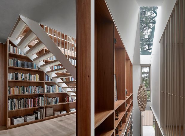 Interior view of a narrow, tall library hall with extensive floor-to-ceiling dark timber shelving, a sculptural white open-riser staircase, and a high skylit void. Bookshelves continue along both sides, with vertical timber screening on the right and glimpses of mature trees through the tall glazing. The space has a refined contemporary architectural character with warm timber joinery and abundant natural light.