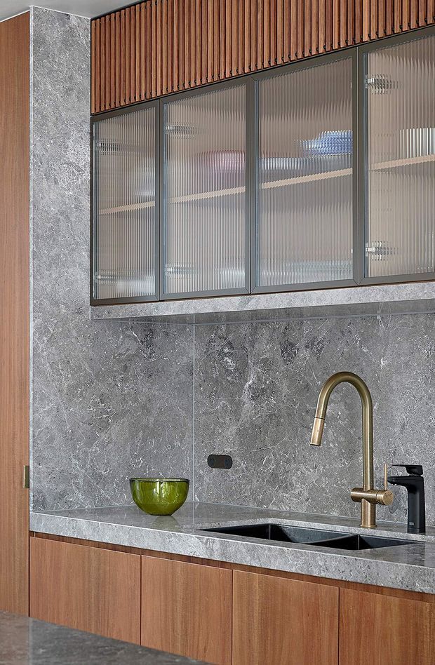 Close-up view of a contemporary kitchen featuring dark grey marble-look wall cladding and benchtops, timber veneer lower cabinetry, vertically fluted translucent-glass upper cabinet doors in dark metal frames, and a brass gooseneck tap over a double-bowl undermount sink. The composition focuses on the kitchen joinery and splashback, with a small green bowl as an accent.