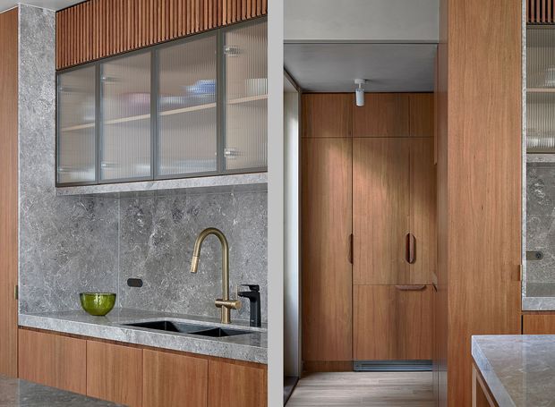 A refined contemporary kitchen interior featuring warm vertical timber cabinetry and slatted timber detailing, a grey veined stone splashback and benchtop, dark undermount sink, and brushed brass gooseneck tap. Ribbed glass-fronted upper cabinetry provides translucent storage. At right, a timber-lined passage or concealed storage area is visible beside the kitchen.