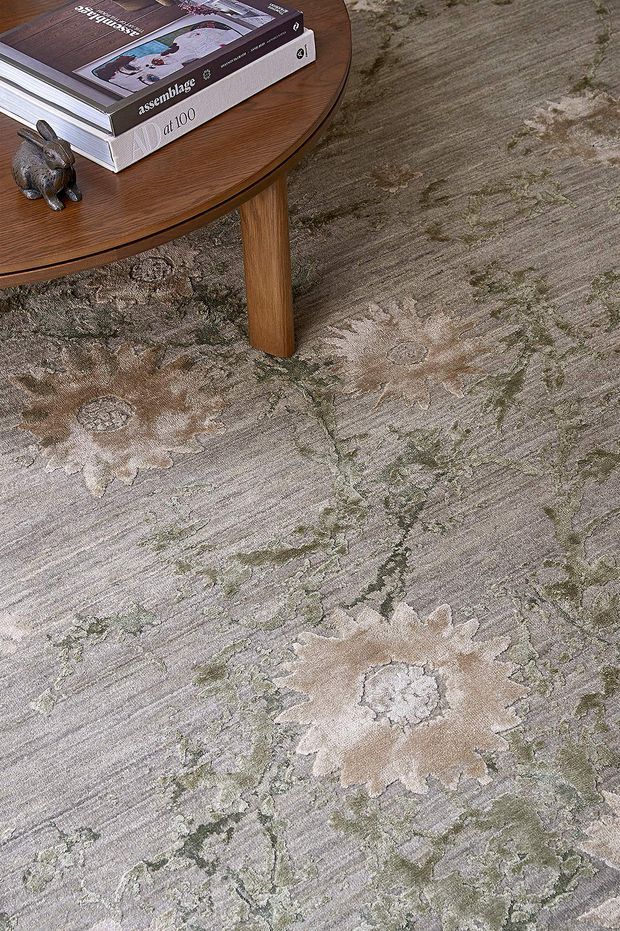 Close-up interior styling image focused on a large patterned carpet or area rug in muted grey, beige, taupe and sage tones. A round mid-brown timber coffee table with a slim edge and central pedestal-style leg sits on the rug, holding stacked design books and a small decorative rabbit. The image primarily showcases the rug's floral and abstract textured pattern.