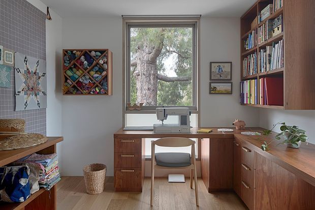 A compact, warm home office and sewing workspace with a built-in timber desk, cabinetry, and wall-mounted bookcases. A sewing machine sits beneath a large window overlooking a tree, while colourful craft materials, framed artwork, textiles, and a potted plant add character. The room has pale walls, timber flooring, and rich mid-century-inspired wood joinery.