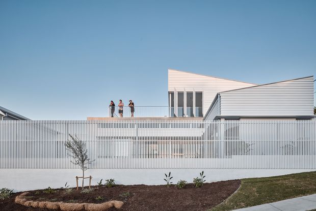 Exterior view of a contemporary, multi-level building with crisp white horizontal cladding and a tall, semi-transparent vertical slat fence enclosing the foreground. A rooftop terrace with three people is visible, along with angular roof forms, extensive glazing, a landscaped planting bed, and a paved walkway under a clear blue sky.