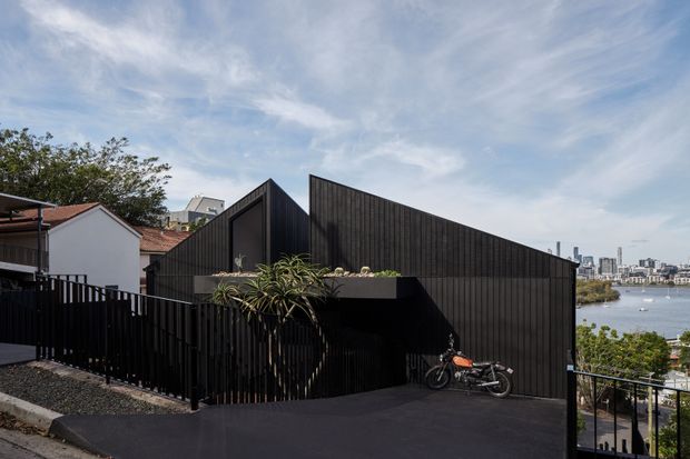 Exterior view of a compact contemporary building with a dramatic angular, gabled form clad in dark vertical metal panels. A black-finished driveway and fence occupy the foreground, with neighbouring houses to the left and a body of water with dense urban development visible beyond the site. A small planted balcony or roof garden projects from the dark facade.