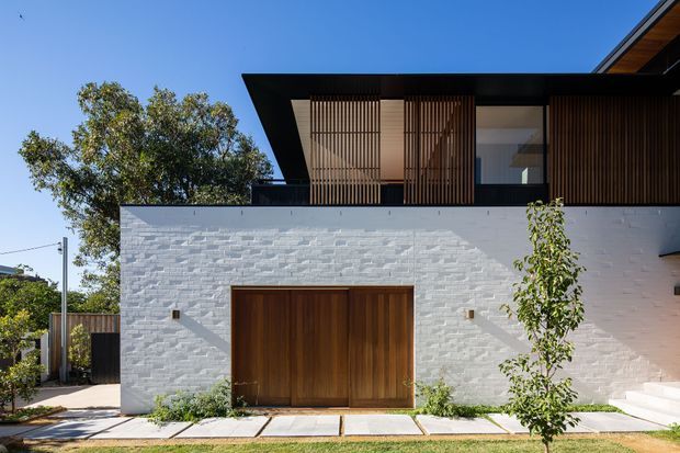 Exterior view of a contemporary two-storey residence with a white painted brick lower facade, broad timber garage door, dark upper cladding, and vertical timber screening around the upper-level glazing. The home has a strong horizontal roofline and crisp geometric composition, set behind a narrow landscaped verge.
