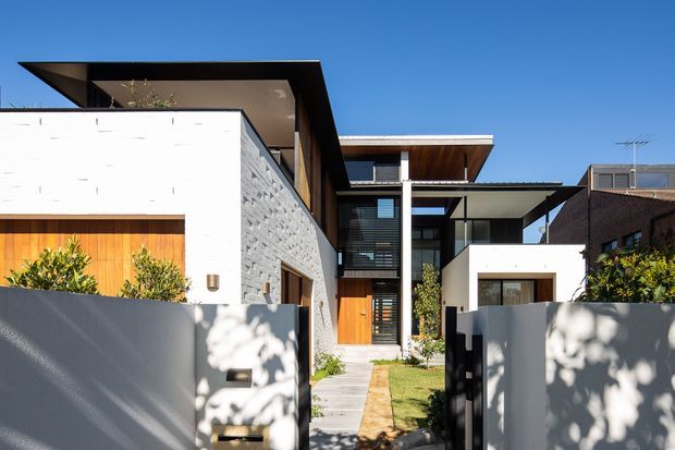 Contemporary multi-level residential facade viewed from the street, featuring crisp white rendered walls, exposed grey masonry, warm timber-clad entry elements, dark roof planes, projecting eaves, and extensive glazing. A gated front path leads through a small landscaped forecourt to the recessed entrance.