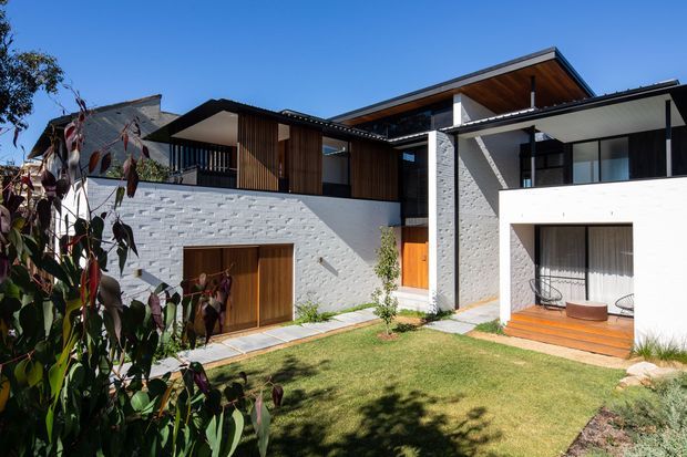 Exterior view of a contemporary multi-level residence with white textured masonry walls, dark-framed glazing, warm timber cladding and soffits, and flat or low-pitched roof planes. The building forms a sheltered courtyard-like lawn with a timber-decked covered outdoor sitting area and integrated garage or entry doors. Landscaping and mature planting frame the foreground.