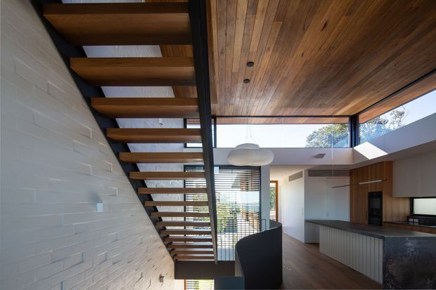 Contemporary open-plan kitchen and living area viewed beneath a dramatic open-tread timber staircase. The space features a warm timber-lined ceiling, exposed white-painted brick wall, dark structural steel, extensive glazing, timber cabinetry, and a dark stone kitchen island. A dining pendant and the edge of a dining chair are visible in the foreground.
