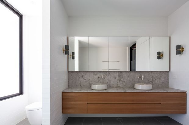 Bright contemporary double-vanity bathroom with a long timber floating vanity, two white vessel basins, mirrored wall cabinets, a mottled grey stone backsplash, white tiled walls, dark floor tiles, and black-and-brass wall sconces. A toilet and large window are visible at the left.