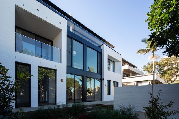 Exterior view of a contemporary multi-storey residence with a white rendered or painted masonry facade, strong black-framed floor-to-ceiling glazing, glass balcony balustrades, recessed balconies, and a flat roofline. Landscaping and neighbouring residential structures are visible around the property.