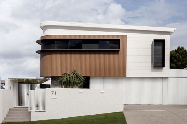 Street-facing view of a contemporary two-storey residence with a curved roofline, white horizontal cladding, warm vertical timber-look wall cladding, dark ribbon windows, and a projecting dark-framed window. A white rendered boundary wall, vertical metal fencing, gated entry, driveway, and minimal front landscaping complete the facade.