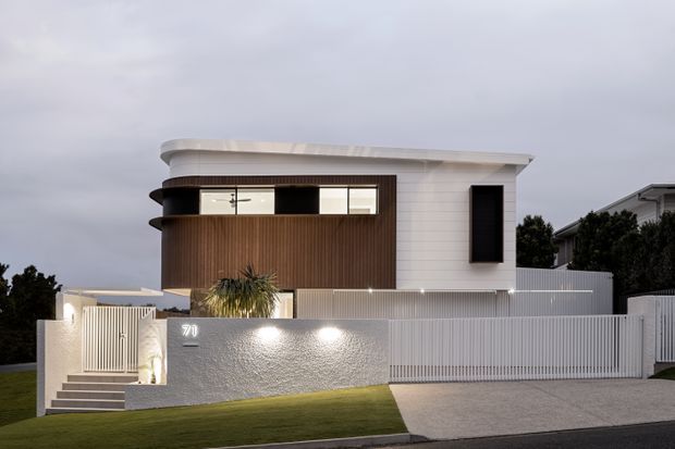 Contemporary two-storey residential facade photographed from the street at dusk. The house features a curved corner balcony and upper roofline, white horizontal cladding, dark vertical timber-look wall cladding, dark-framed windows, a rendered boundary wall, vertical metal fencing, front steps, and a concrete driveway.