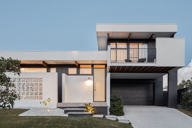 Contemporary two-storey residential facade with a clean white and dark grey rendered exterior, projecting upper balcony, vertical metal balustrades, flat rooflines, timber-toned soffit details, and a recessed double garage. A decorative breeze-block screen and landscaped front garden soften the minimalist geometric composition.