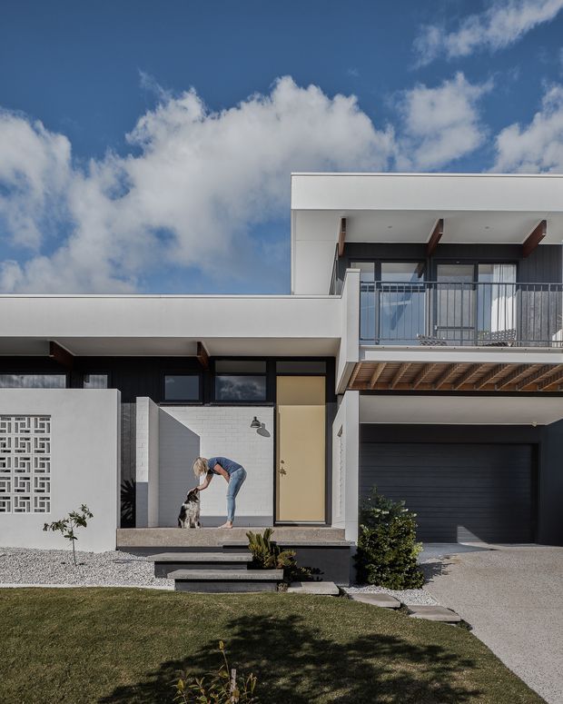 Street-facing view of a contemporary two-storey house with a crisp white rendered facade, dark cladding, flat roof forms, projecting balconies, exposed timber soffit structure, and a dark garage door. The entry is approached by stepped concrete pavers through a sparse landscaped front yard, with a decorative breeze-block screen beside the entry. A person and dog are present near the doorway.