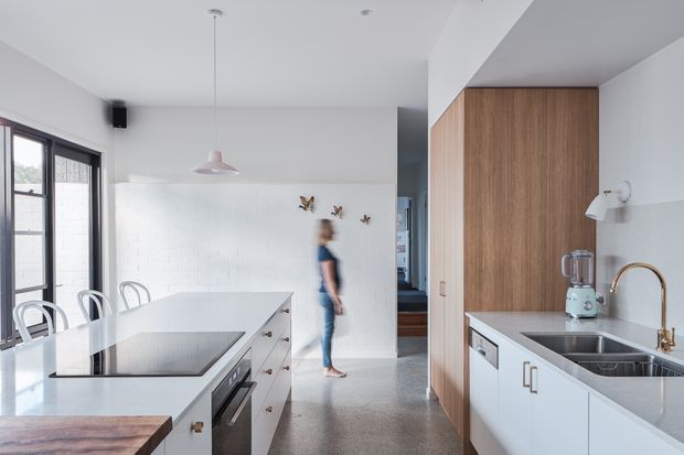 Bright contemporary kitchen with a long white island, integrated black cooktop, white cabinetry, timber-lined tall joinery, stainless-steel sink and warm brass tapware. Large black-framed windows illuminate a polished concrete or terrazzo-like floor, while a white painted brick feature wall and pendant lighting add texture. The open layout includes a glimpse toward an adjoining living area.
