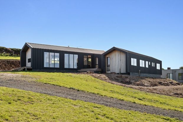 Single-storey contemporary house on a sloping grassy site, composed of dark corrugated-clad wings with a prominent pitched gable section. Large glazed openings and vertical timber-toned cladding add contrast to the dark exterior.