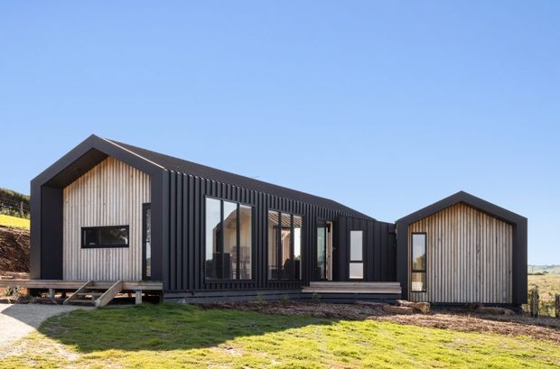 Exterior view of a contemporary single-storey modular or prefabricated home composed of two gabled volumes connected by a dark-clad central section. The facade combines black ribbed vertical cladding with pale timber-look vertical boards, large glazed openings, and a raised deck and entry steps. The building sits in an open rural or semi-rural landscape under a clear blue sky.