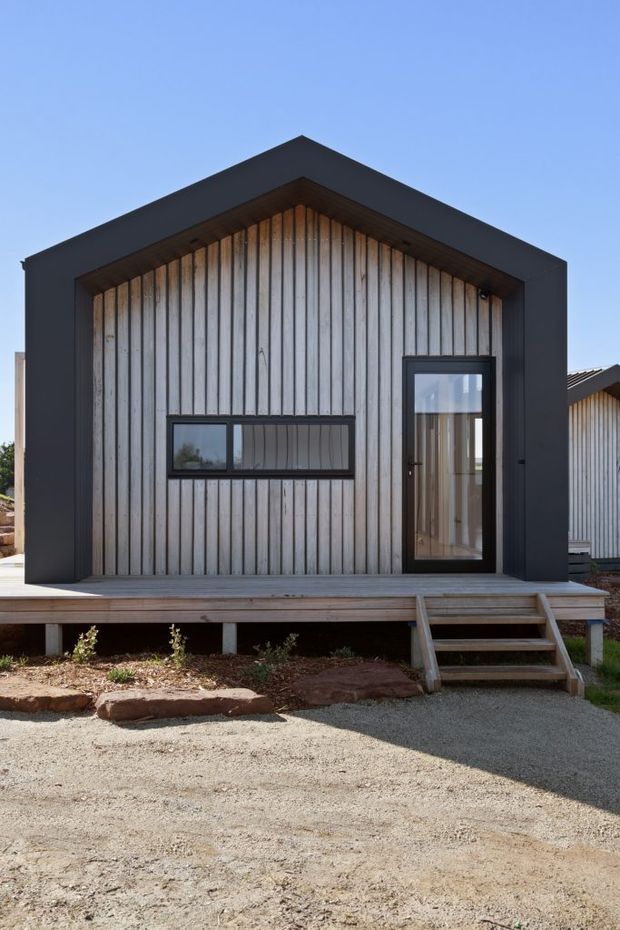Front exterior view of a compact raised single-storey building with a steep gabled roof form, vertical weatherboard cladding, a broad dark-painted geometric facade frame, and dark-framed glazing. A timber deck and small stair lead to the glazed entry door. The building sits on a gravelled site with simple landscaping.