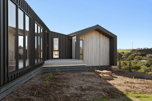 Exterior view of a contemporary multi-wing house on a sloping, landscaped site. The building features dark vertical cladding, extensive floor-to-ceiling glazing, a light timber-lined gable facade, and a raised timber deck with steps. Rolling green landscape is visible beyond the property.